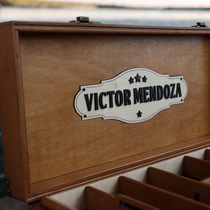 May include: A wooden box with a hinged lid and a sign that reads "VICTOR MENDOZA" with stars. The box has multiple compartments inside.