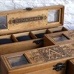 May include: A wooden box with a glass top and a compartment for storing watches. The box is engraved with the words "Zeitvertreiber" and a decorative design.