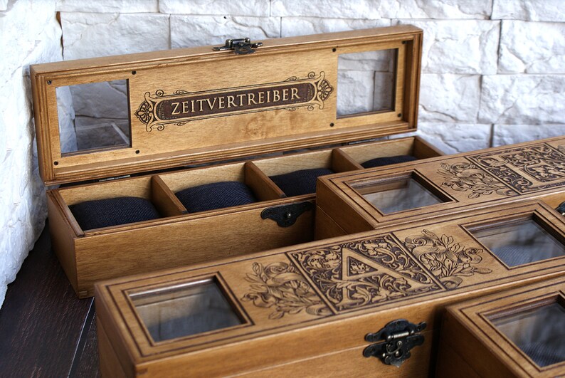 May include: A wooden box with a glass top and a compartment for storing watches. The box is engraved with the words "Zeitvertreiber" and a decorative design.