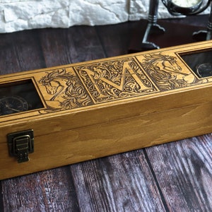 May include: A wooden watch box with a hinged lid and two clear glass windows. The box is engraved with a stylized letter "M" surrounded by floral designs and two horses. The box has two brass clasps.