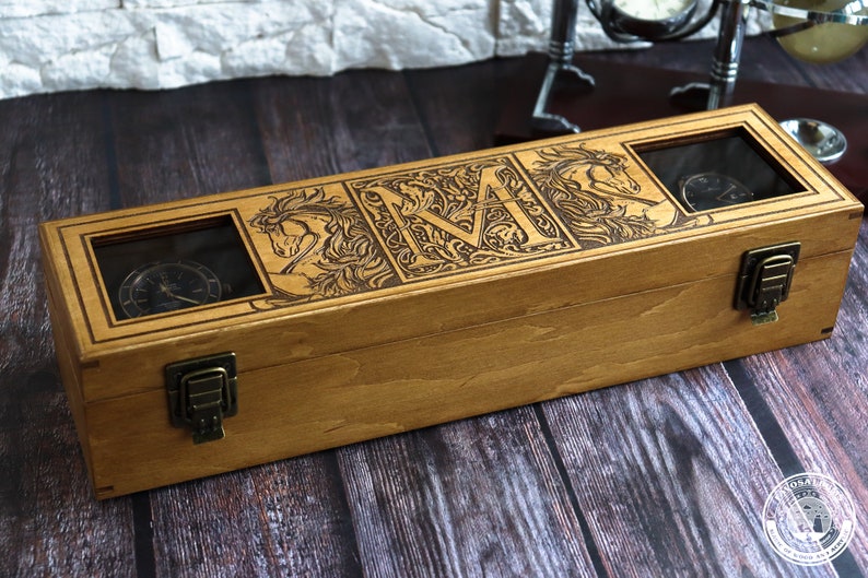 May include: A wooden watch box with a hinged lid and two clear glass windows. The box is engraved with a stylized letter "M" surrounded by floral designs and two horses. The box has two brass clasps.