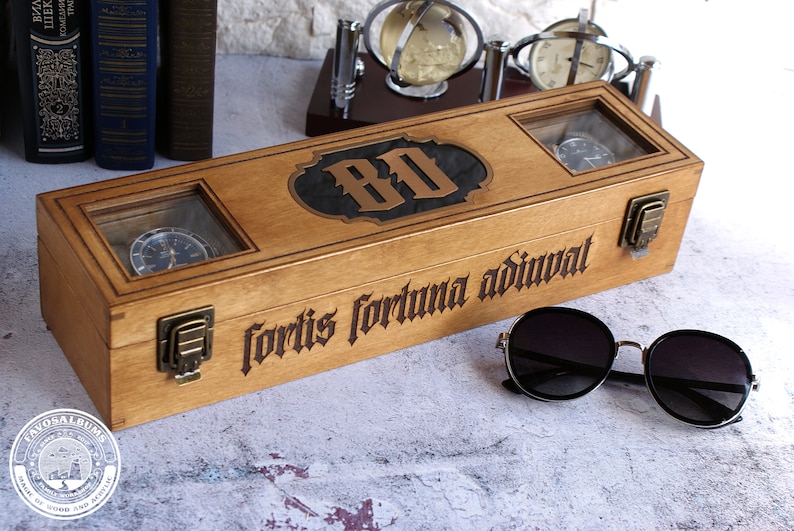 Peut inclure: Une bo&icirc;te &agrave; montres rectangulaire en bois avec deux fen&ecirc;tres en verre, des loquets en laiton et le texte grav&eacute; "fortis fortuna adiuvat" sur le devant. Le couvercle pr&eacute;sente une incrustation sombre centrale avec les lettres "BO".