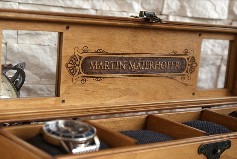 Pu&ograve; includere: Una scatola porta orologi in legno con un coperchio in vetro e un'iscrizione personalizzata che recita "MARTIN MAIERHOFER". La scatola ha un interno in velluto con scomparti per riporre gli orologi.