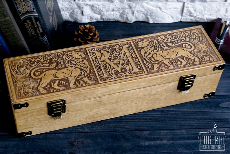 May include: A wooden box with a hinged lid, featuring intricate carvings of lions and a stylized letter "M". The box has brass hinges and clasps.