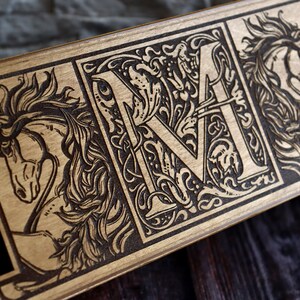 May include: A wooden watch box with a black velvet interior and three compartments. The box is decorated with a laser-engraved design featuring a horse, a letter "M" with floral accents, and another horse. The box has a circular logo on the bottom right corner that reads "Favosalums Made of Wood and Acrylic".