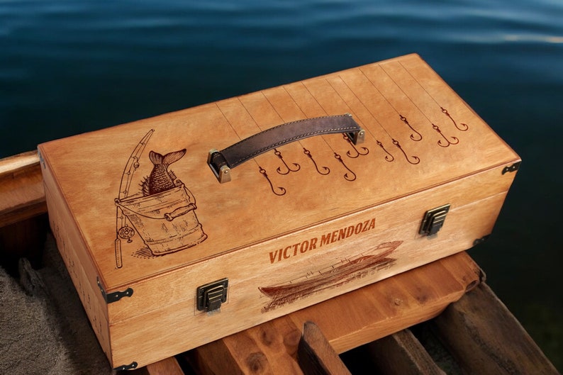 May include: A wooden fishing tackle box with a brown leather handle. The lid is engraved with a fish in a bucket, a fishing rod, and the text "VICTOR MENDOZA".