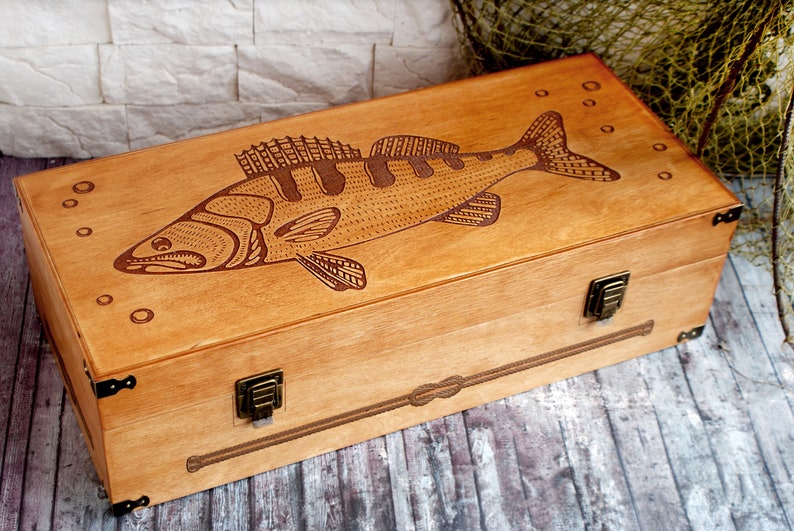 May include: A wooden box with a hinged lid and brass clasps. The top of the box is decorated with an engraved image of a fish, possibly a walleye or pike, with bubbles around it.