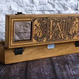 May include: A wooden box with a hinged lid, featuring a decorative design with the letters "LAN" and floral motifs. The box has two glass windows and a label with the text "www.zavod.co.uk 775710".