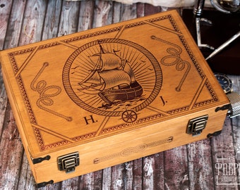Puede incluir: Una caja de madera con temática náutica. La tapa está grabada con un barco, una cuerda y una brújula. La caja tiene dos cierres de latón y las iniciales "H. J." están grabadas en la tapa.