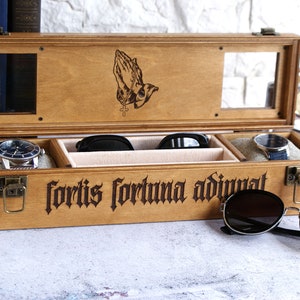 Peut inclure: Une bo&icirc;te de rangement rectangulaire en bois avec un couvercle &agrave; charni&egrave;re, orn&eacute;e d'une gravure laser repr&eacute;sentant des mains en pri&egrave;re et le texte latin fortis fortuna adiuvat. L'int&eacute;rieur comporte des compartiments pour deux montres.