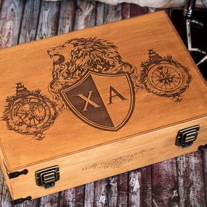 K&ouml;nnte beinhalten: Eine Holzkiste mit einem L&ouml;wen, einem Kompass und einem Schild, der auf den Deckel graviert ist. Der Schild tr&auml;gt die Initialen "XA".