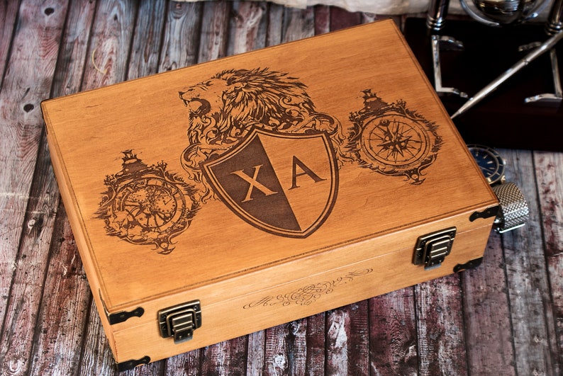 K&ouml;nnte beinhalten: Eine Holzkiste mit einem L&ouml;wen, einem Kompass und einem Schild, der auf den Deckel graviert ist. Der Schild tr&auml;gt die Initialen "XA".