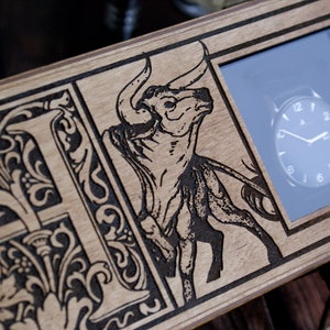 May include: A wooden box with a black and white engraved design of a bull and a decorative letter "F". The box has a glass window that displays a watch with black hands and a white face.