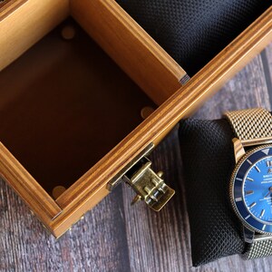 May include: A blue watch with a silver band and a blue face. The watch is sitting on a black cushion inside a wooden box.