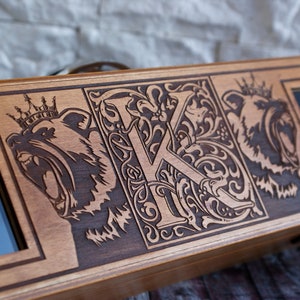 May include: A wooden box with a hinged lid, featuring an intricate design of a crowned bear, a stylized letter "K", and another crowned bear. The box has two small rectangular openings on either side.