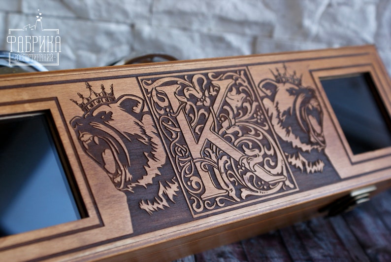 May include: A wooden box with a hinged lid, featuring an intricate design of a crowned bear, a stylized letter "K", and another crowned bear. The box has two small rectangular openings on either side.