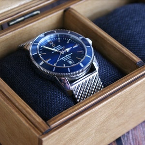 May include: A silver watch with a blue face and a mesh metal band is displayed in a wooden box with a blue velvet lining.
