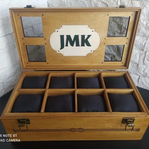 May include: Wooden watch box with eight compartments, each with a black cushion. The box has a light brown finish, a hinged lid, and a decorative clasp. The lid features the initials "JMK" in green on a white background.