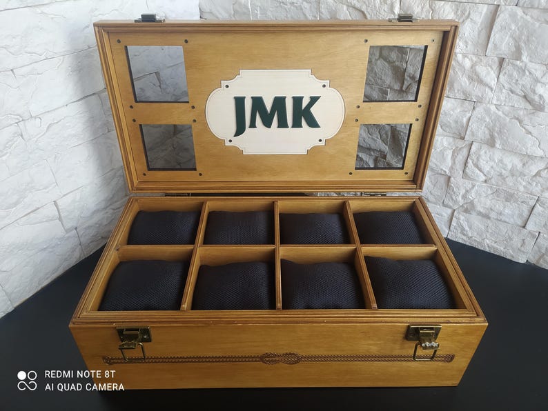 May include: Wooden watch box with eight compartments, each with a black cushion. The box has a light brown finish, a hinged lid, and a decorative clasp. The lid features the initials "JMK" in green on a white background.