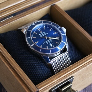 K&ouml;nnte beinhalten: Eine silberne und blaue Breitling Superocean Uhr mit einem Mesh-Armband in einer Holzkiste mit schwarzem Samtbezug.