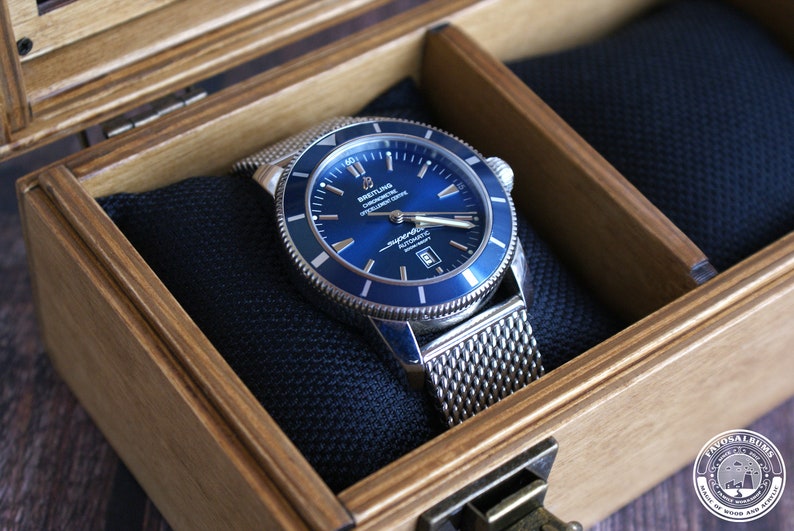 K&ouml;nnte beinhalten: Eine silberne und blaue Breitling Superocean Uhr mit einem Mesh-Armband in einer Holzkiste mit schwarzem Samtbezug.
