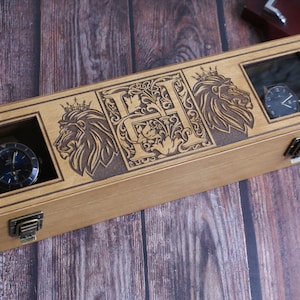 Puede incluir: Una caja de madera para relojes con dos compartimentos para relojes y un diseño de cabeza de león con una corona a cada lado. La caja está grabada con la letra "H" en una fuente decorativa.