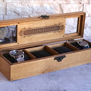 May include: A wooden watch box with a glass top and three compartments. The box is engraved with the name "VIVIEN BRISSAT".
