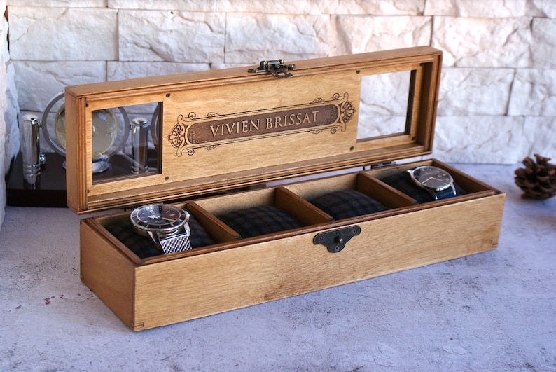 May include: A wooden watch box with a glass top and three compartments. The box is engraved with the name "VIVIEN BRISSAT".