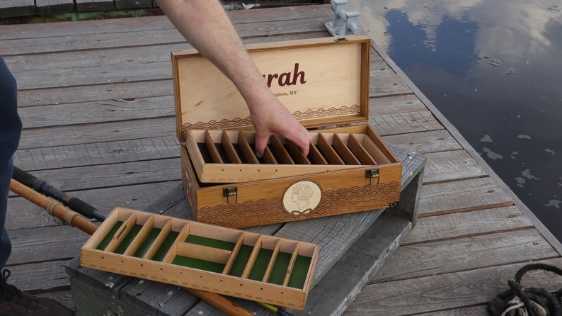 May include: A rectangular wooden storage box with a hinged lid featuring the text "rah" and "ington, NY". The box has a removable tray with green felt lining and multiple dividers. The exterior features a circular emblem and wave patterns.