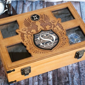 May include: A wooden watch box with a glass top and a decorative engraved design featuring a shield with the letter "S" and two lions. The box has two brass clasps and is ideal for storing and displaying watches.