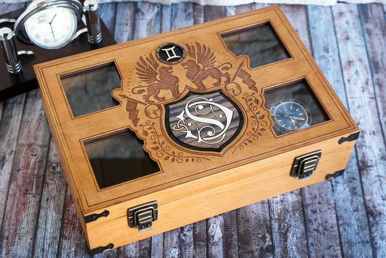 May include: A wooden watch box with a glass top and a decorative engraved design featuring a shield with the letter "S" and two lions. The box has two brass clasps and is ideal for storing and displaying watches.