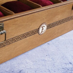 May include: A wooden watch box with a brown finish and a decorative inlay. The box has a brass latch and a circular inlay with the letter "E" in the center.