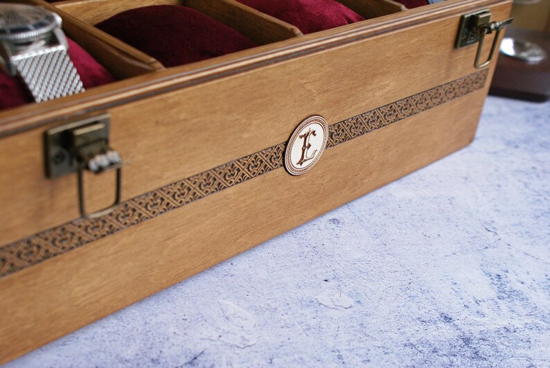 May include: A wooden watch box with a brown finish and a decorative inlay. The box has a brass latch and a circular inlay with the letter "E" in the center.