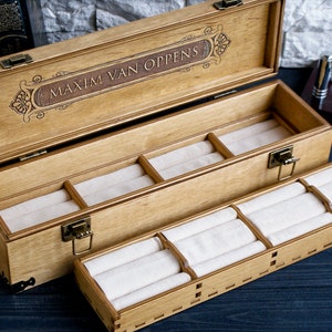 May include: A wooden jewelry box with a pull-out drawer. The top of the box is engraved with the name "Maxim Van Oppens". The box has a brown stain and is lined with white velvet.