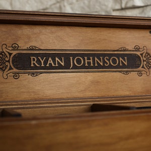 May include: A wooden box with a personalized inscription that reads "RYAN JOHNSON" in a decorative font. The box has a dark brown stain and a clear glass window on each side.