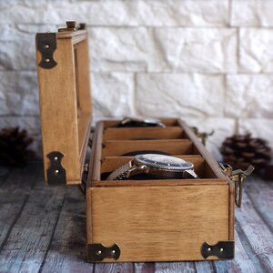 May include: A wooden watch box with a hinged lid and brass accents. The box has three compartments for storing watches. The box is open and a silver watch is visible in one of the compartments.