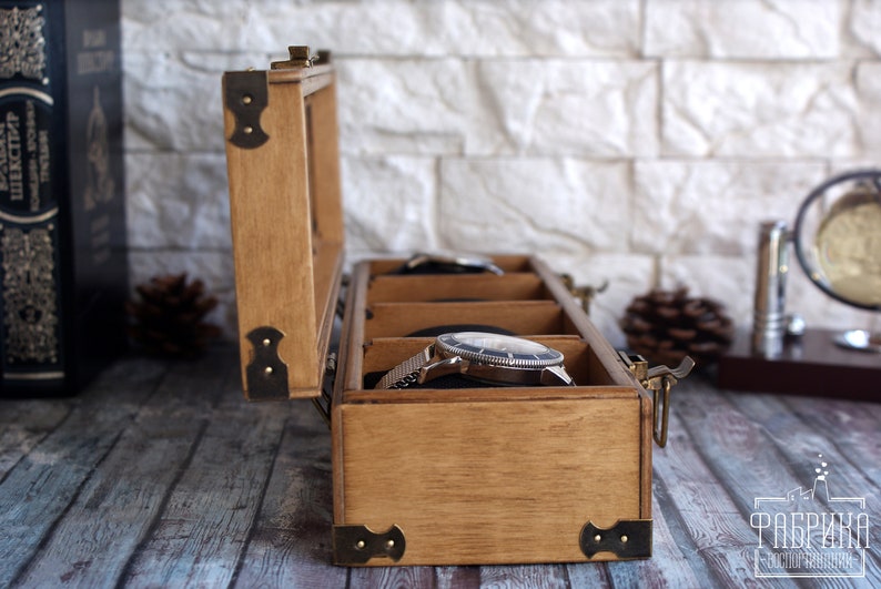 May include: A wooden watch box with a hinged lid and brass accents. The box has three compartments for storing watches. The box is open and a silver watch is visible in one of the compartments.