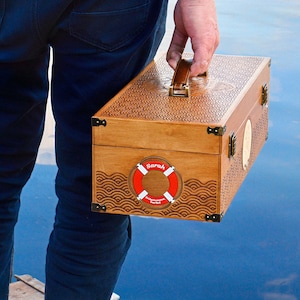 May include: A rectangular wooden storage box with a brown finish, featuring an intricate laser-engraved wave pattern on the lid and sides. The box has a leather handle, metal corner accents, and a red and white lifebuoy graphic with the name Sarah.