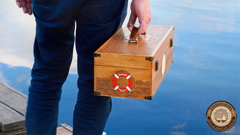 May include: A rectangular wooden storage box with a brown finish, featuring an intricate laser-engraved wave pattern on the lid and sides. The box has a leather handle, metal corner accents, and a red and white lifebuoy graphic with the name Sarah.