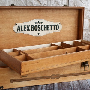 May include: Wooden storage box with a hinged lid and a removable tray. The box is light brown with a decorative design and the name "ALEX BOSCHETTO" on the lid. The interior has multiple compartments for organizing items.