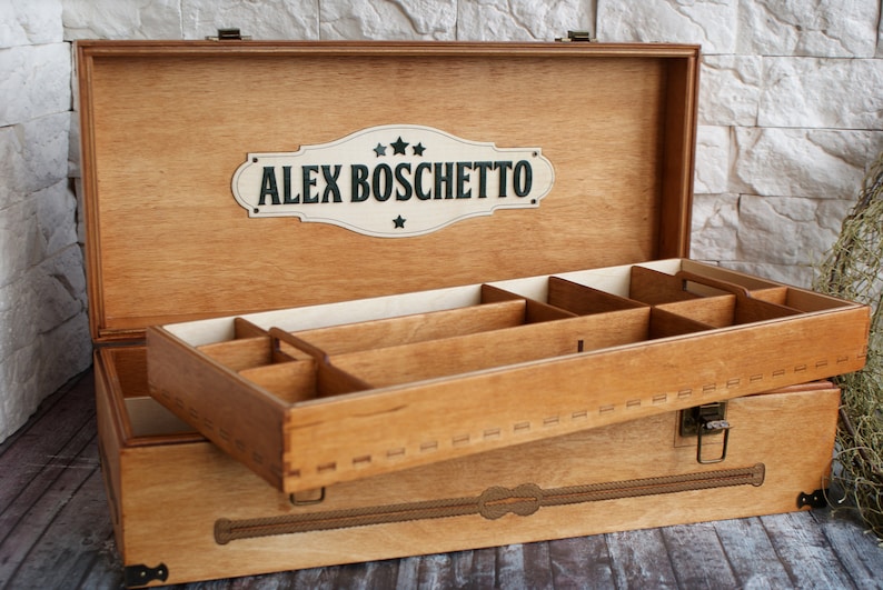May include: Wooden storage box with a hinged lid and a removable tray. The box is light brown with a decorative design and the name "ALEX BOSCHETTO" on the lid. The interior has multiple compartments for organizing items.