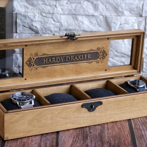 May include: A wooden watch box with a glass top and four compartments. The box is engraved with the name "Hardy Draxler".