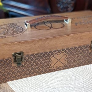 May include: A wooden fishing tackle box with a brown leather handle and metal clasps. The box features engraved designs, including a compass rose and a fish hook symbol. The lower section has a scale-like pattern with a four-leaf clover.
