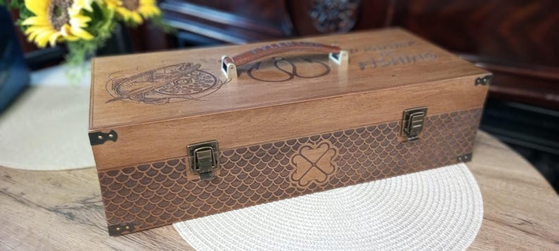 May include: A wooden fishing tackle box with a brown leather handle and metal clasps. The box features engraved designs, including a compass rose and a fish hook symbol. The lower section has a scale-like pattern with a four-leaf clover.