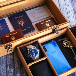 Puede incluir: Una caja de madera con un pasaporte azul, un pasaporte rojo, un documento blanco y un tel&eacute;fono azul. La caja tambi&eacute;n tiene un reloj con una esfera azul y una correa plateada, y un juego de llaves.