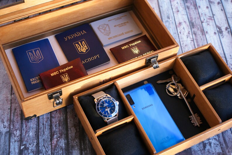 Puede incluir: Una caja de madera con un pasaporte azul, un pasaporte rojo, un documento blanco y un tel&eacute;fono azul. La caja tambi&eacute;n tiene un reloj con una esfera azul y una correa plateada, y un juego de llaves.