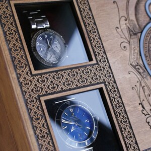 May include: A wooden box with two watch display compartments. The box has an intricate design and a clock face on the front. The watches are both silver with black faces.