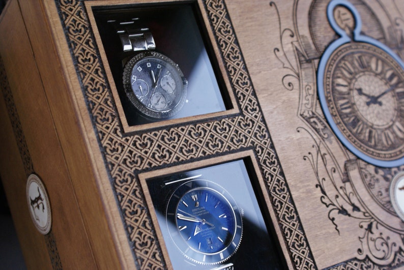 May include: A wooden box with two watch display compartments. The box has an intricate design and a clock face on the front. The watches are both silver with black faces.