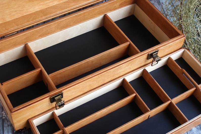 May include: Open wooden box with multiple compartments. The box is made of light brown wood with a black interior and a brass-colored clasp. The compartments are divided by wooden dividers, creating various storage spaces. The box is designed for organizing small items.