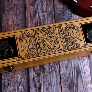May include: A wooden box with a hinged lid, engraved with a decorative floral design and the letter "M". The box has three compartments, each with a cutout for a watch. Two watches are visible, one with a black face and the other with a silver face.